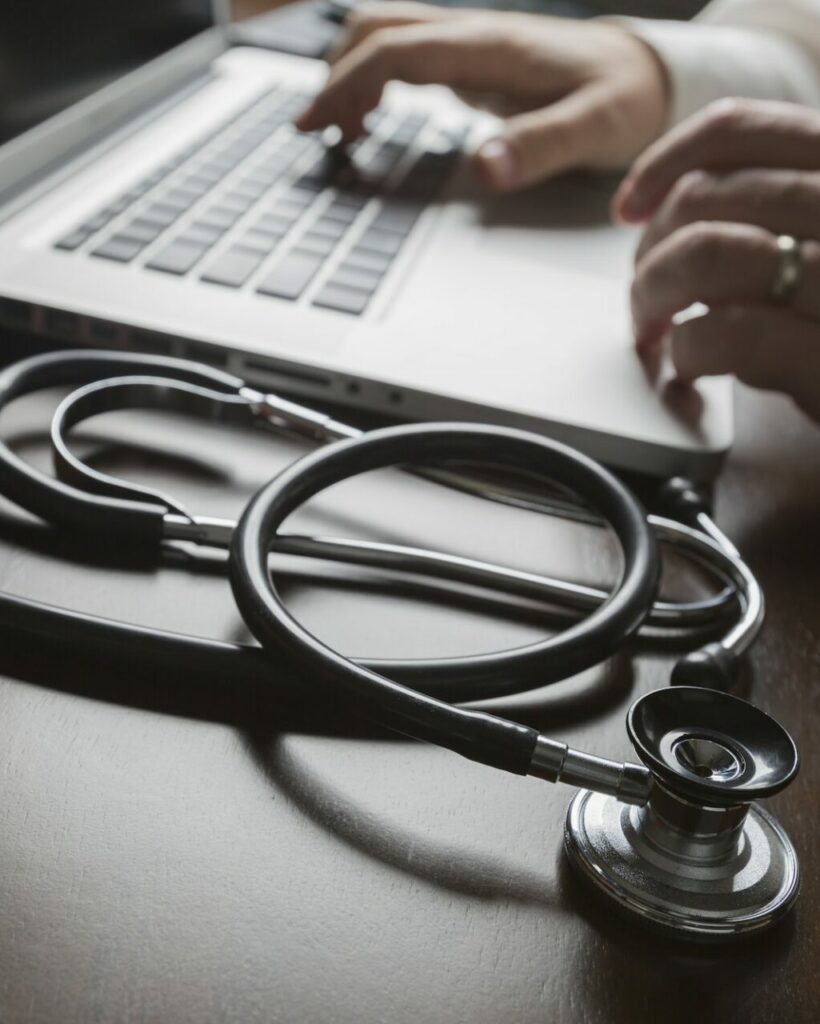 Medical Stethoscope Resting on Desk As Male Hands Type on Computer Keyboard.